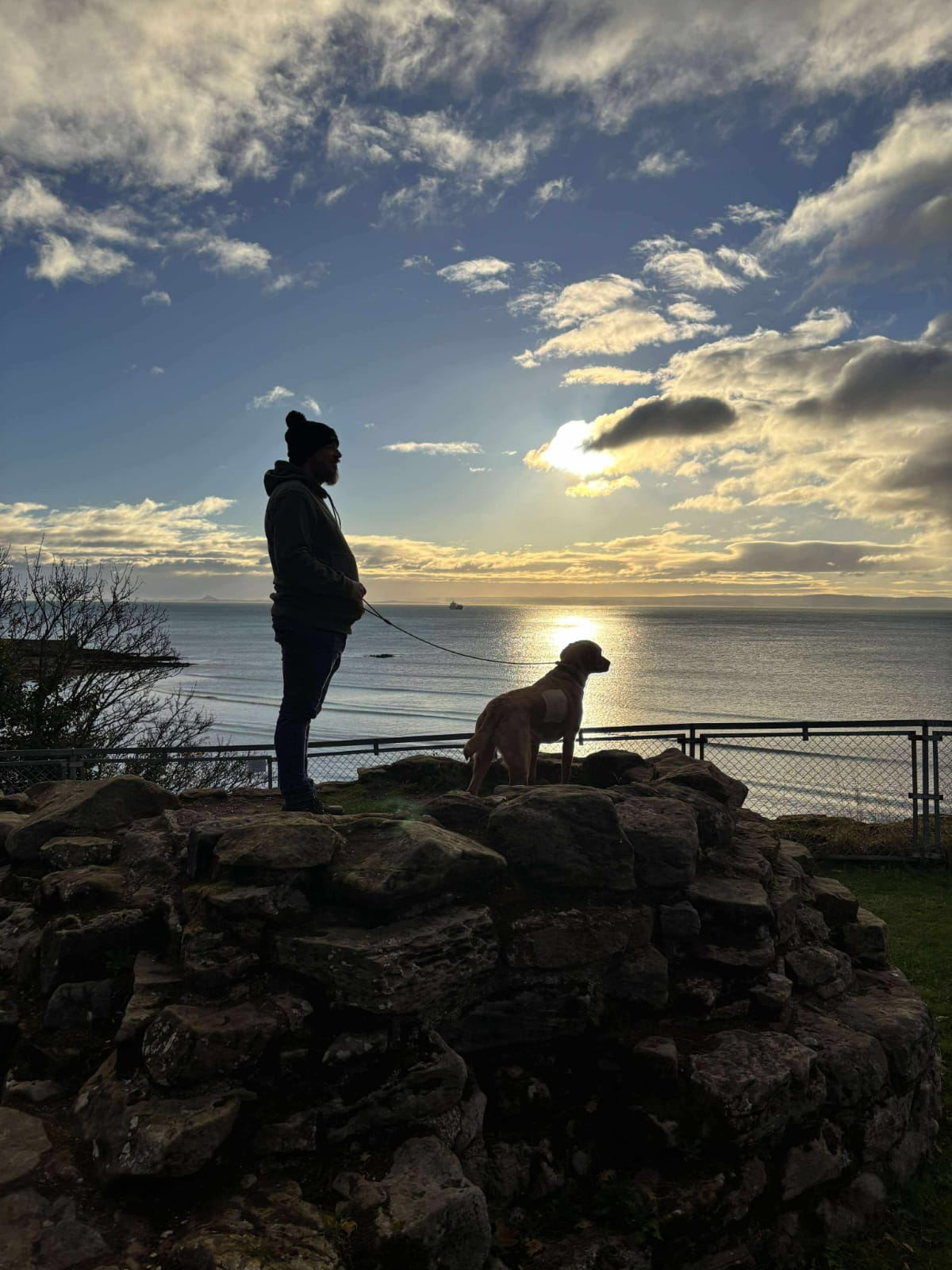 Blair standing on rocks overlooking the sea with a dog at sunset